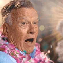 An elderly white man wearing flower leis looking shocked