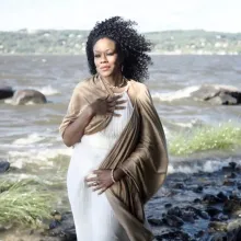 A Black femme in a white dress looks tranquilly toward the camera, as in Botticelli’s Birth of Venus, at the bank of a river.