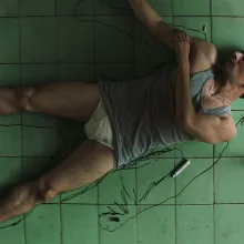 A man with brown skin and short hair, wearing a tank top and underwear, traces himself on the floor.