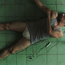 A man with brown skin and short hair, wearing a tank top and underwear, traces himself on the floor.