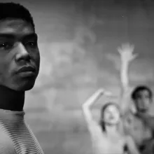Black and white image of a black man with short dark hair looking into the camera. In the background three dancers strike a pose.