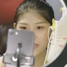 An asian woman with black hair holds up a white tennis shoe in front of a camera and ring light.