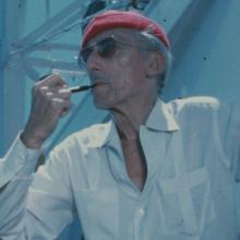 An older white man wearing a red cap, white shirt and sunglasses. He is smoking a pipe.