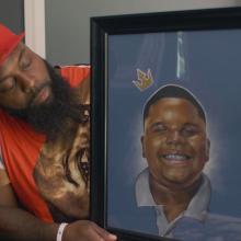 A Black man with a long black beard wearing a red baseball cap and shirt holds a framed painting of a Black boy with a crown on his head.