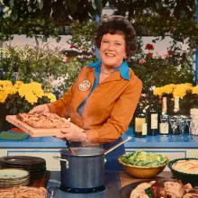 A woman with white skin and curly brown hair. She holds a pan with food on it.