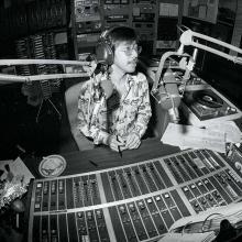 A black and white photo of an asian man in a patterned button up. He sits in a recording studio.