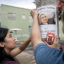 A Latinx woman with black hair and someone off screen hang a poster that reads "MISSING PERSON." It has a picture of a Latinx man with short black hair on it.