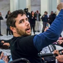 A white man with brown hair and facial hair raises his fist in the air. He is in a wheelchair.
