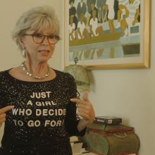 An older white woman with gray hair and black glasses. She is pointing to her shirt which reads "Just A Girl Who Decided To Go For It."