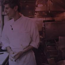 Anthony Bourdain, an older white man with short hair wearing a white chef's coat, stands in a small kitchen.