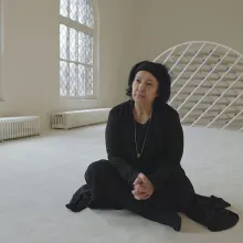 An older woman with brown skin and black hair wearing a black outfit sits on the floor.