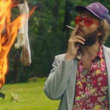 A white man with brown hair and facial hair wearing red shades and a Hawaiian shirt and blazer. He smokes a cigarette and watches socks burn.
