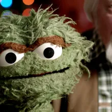 An older white man with gray hair holds up a green, scraggly puppet.