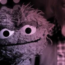 An older white man with gray hair holds up a green, scraggly puppet.