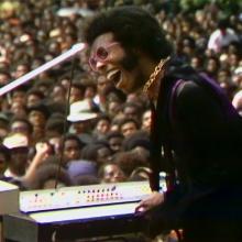 A Black man with black hair wearing pink glasses, a gold necklace and a purple suit. He is playing keyboard to a crowd.