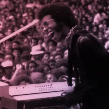 A Black man with black hair wearing pink glasses, a gold necklace and a purple suit. He is playing keyboard to a crowd.