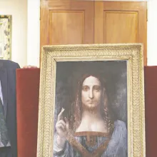 An older white man with black glasses and gray short hair stands next to Salvator Mundi, a painting of a white man with long brown hair pointing upwards.