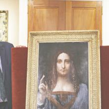 An older white man with black glasses and gray short hair stands next to Salvator Mundi, a painting of a white man with long brown hair pointing upwards.