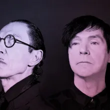 A black and white portrait of two white men both with dark hair. The man on the left has round black glasses and a pencil mustache.