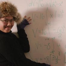 A young Asian student with glasses, wearing a goofy fur hat, motions to a white board with a math formula on it.