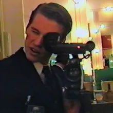A white man with short brown hair holds a camera up to a mirror.