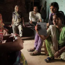 A group of people with brown skin sit in a room. Two women in the front film someone off screen with their phone.