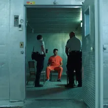 A man in orange jumpsuit is being interrogated by officers