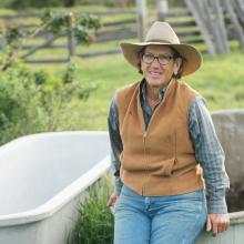 A woman wearing a cowboy hat and vest sitting on a washtub outside in a garden.