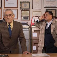 An elderly man in a brown suit and a middle-aged man in trench coat are behind an office desk