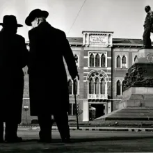 Two men in winter coats and hats in the style of the early 20th century speak to one another closely before a large courtyard with a statue at its center. 