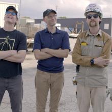 Three Caucasian male are smiling at the camera with their arms interlocked.