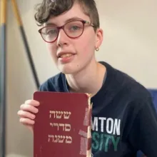 An image of non-binary teen AJ Hack holding his family's copy of the Mishnah, a red book with Hebrew lettering and a taped-together binding. 