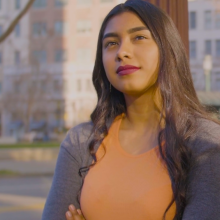 A young Latina woman stands with arms crossed in front of distant buildings