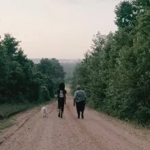 Two women is walking with their white dog on an unpaved road flanked by green green trees.