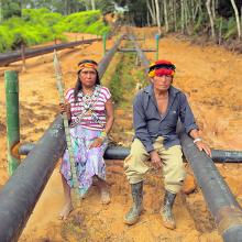 Two indigenous people sitting on black pipes in the Amazonian forest.