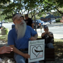 A white man with a beard holds an anti-refugee sign in a residential neighborhood.