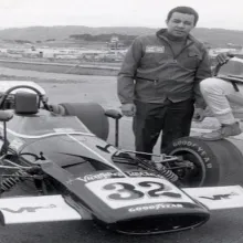 two black male racers stand next to a racecar at a speedway.