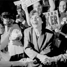 Barbara Boxer is shaking hands with others while supporters hold signs in the background.