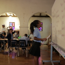 a grade school African American girl in a school uniform looks up at a white board with a paper in her hand in front of the rest of her young classmates.