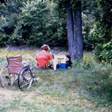 two camp members sit under a tree while attending a disabled persons camp; a wheel chair is focal point of image