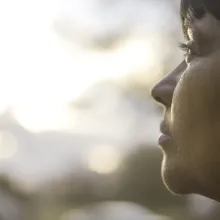 A still from "Commuted": Protagonist Danielle Metz, a middle aged woman, is shown in sharp focus in the foreground, looking out at a sunset. 