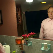 white middle aged male stares at himself in the bathroom mirror wearing pink button up