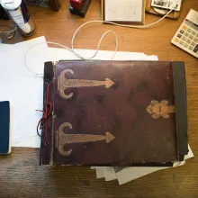 A rustic old notebook of archived documents sits on a desk surrounded by smaller papers and notepads.