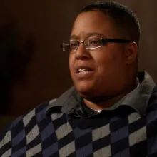 A middle-aged black individual in a patterned sweater sits in front of a muted brown backdrop