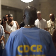 A Black Male Folsom inmate stands in a blue shirt in front of a diverse group of male prisoners.