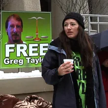 A woman stands in front of a 'Free Greg Taylor' poster.