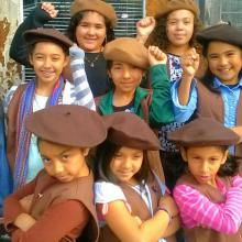 A group of young girls of color stand in brown berets with their fists raised. 