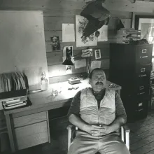 Jim Harrison sits with his hands in his lap in his rugged, outdoorsy 20th-century office. 