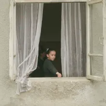 A young girl stares out the open window of a rustic stone building