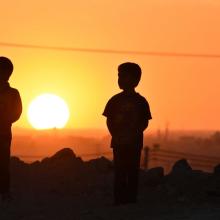 Two silhouetted children stand before the setting sun.